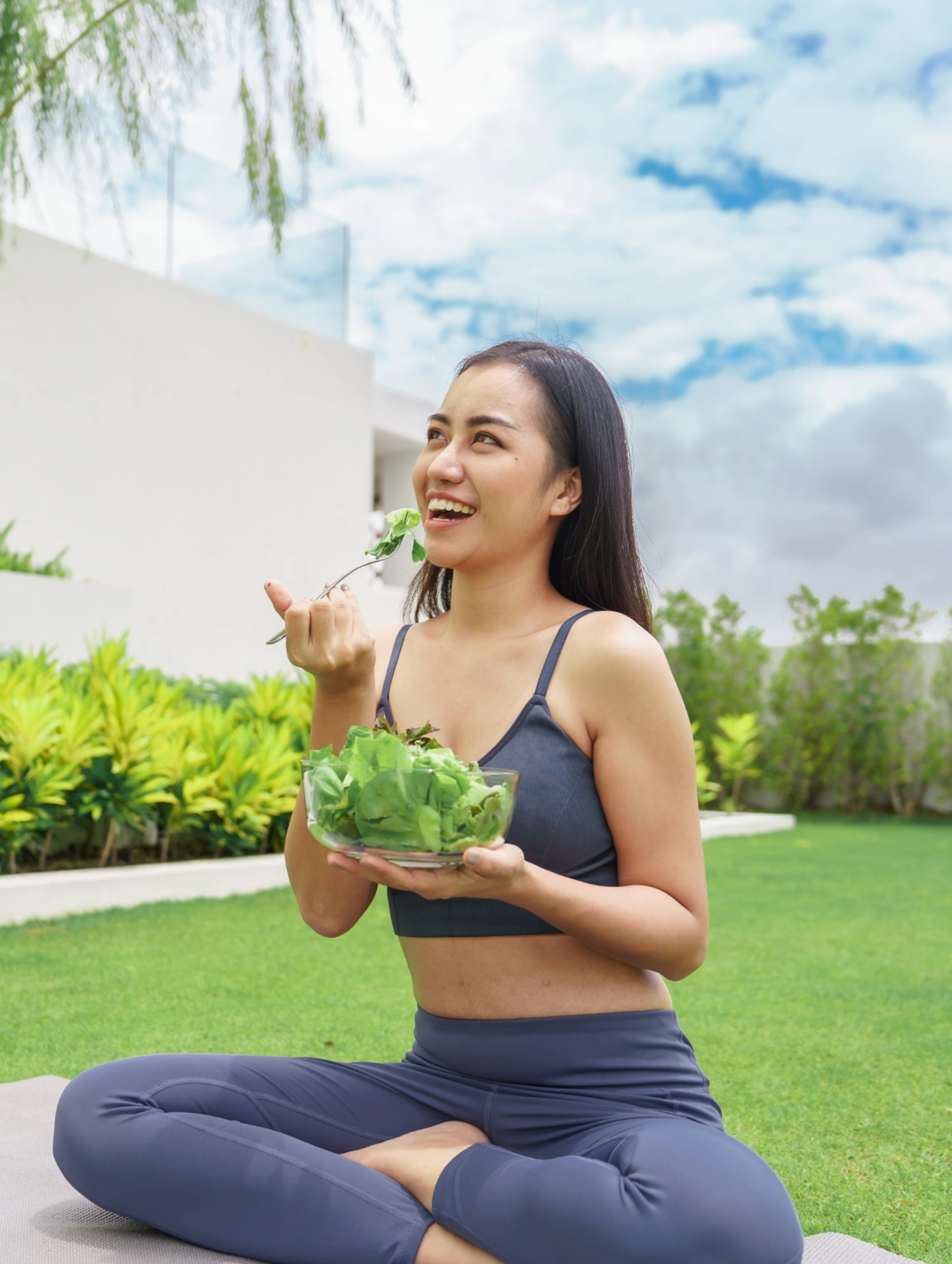 Woman in activewear sitting outdoors on a yoga mat while eating a fresh green salad, healthy lifestyle image representing simple and satisfying snacks for clean eating.