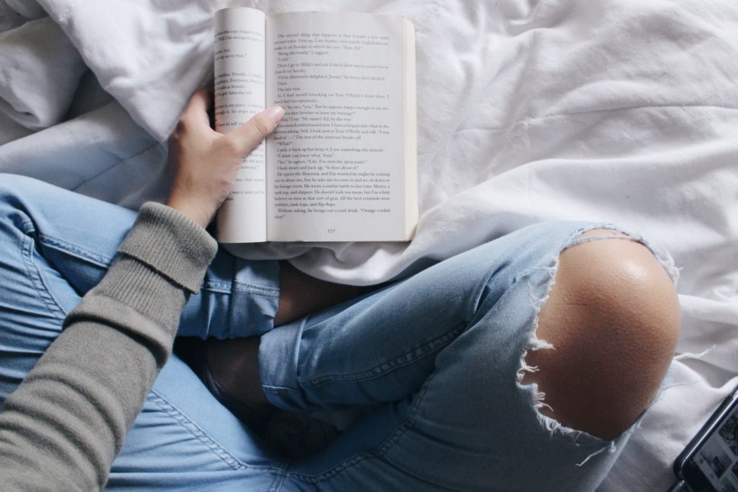 Woman in ripped jeans reading a book on her bed
