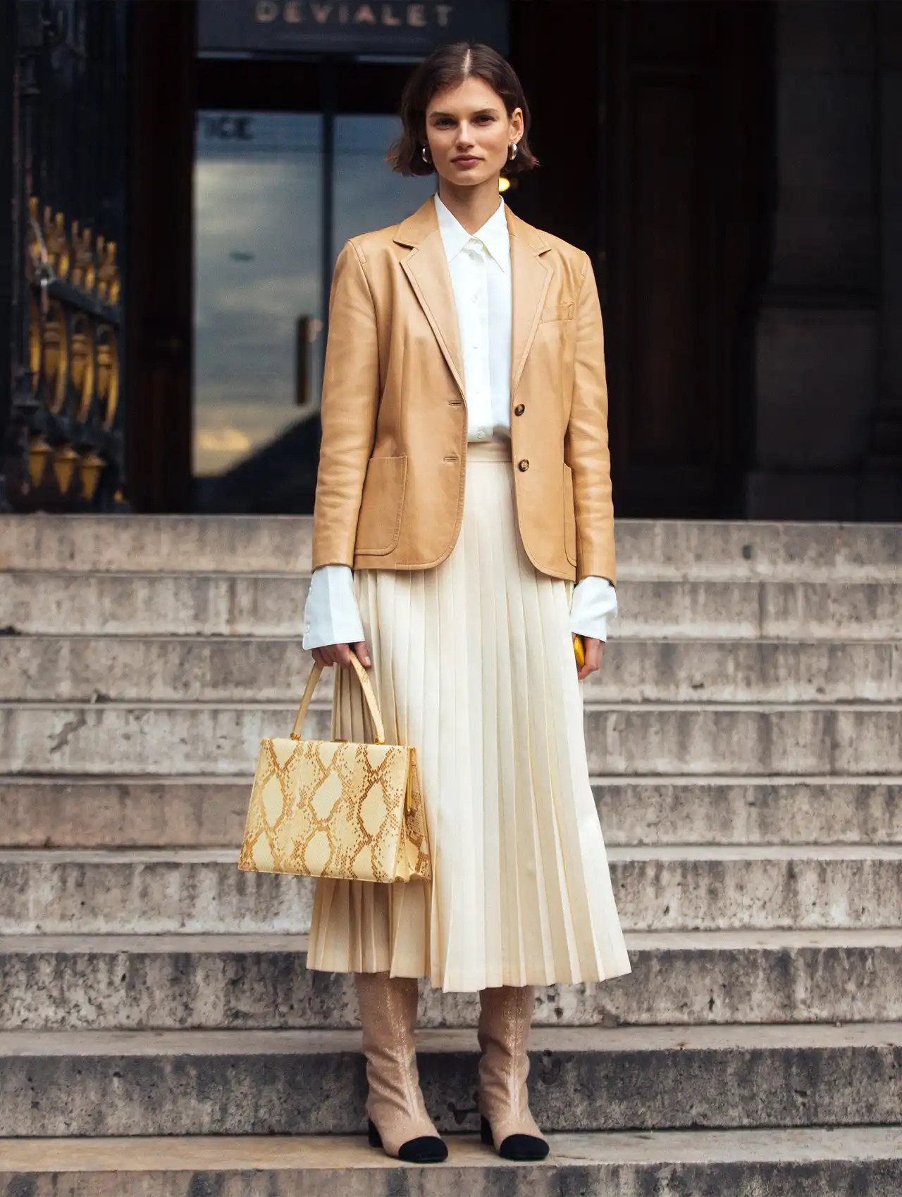 Woman in a beige blazer for women paired with a pleated midi skirt and boots on city steps.