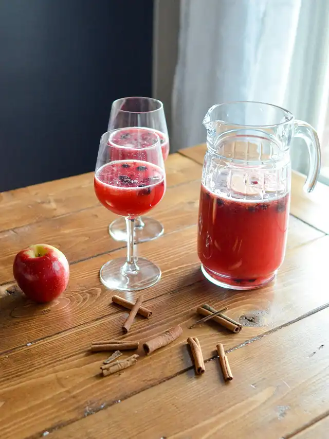 a glass of red liquid next to a pitcher of cinnamon sticks and an apple