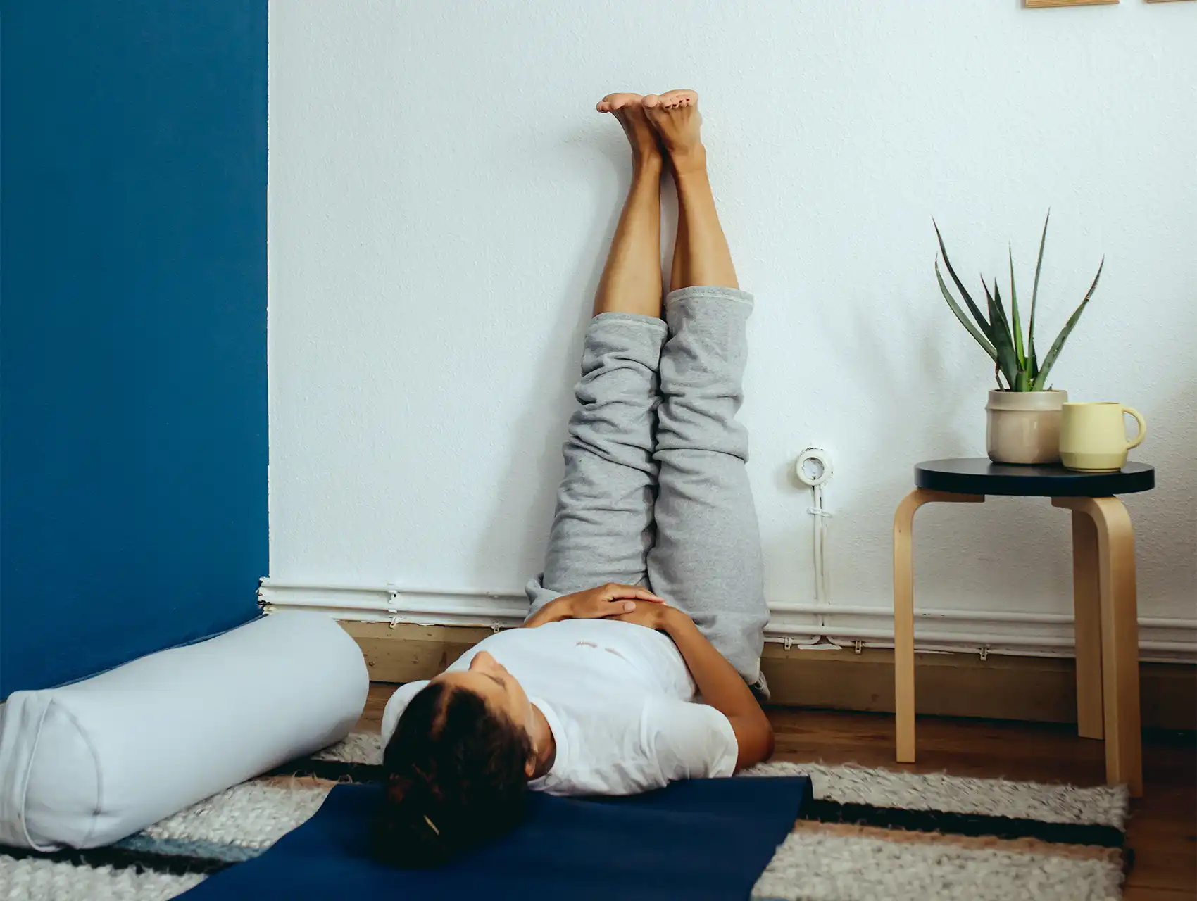 Woman practicing vertical leg extension with white bolster beside her yoga mat with bolsters
