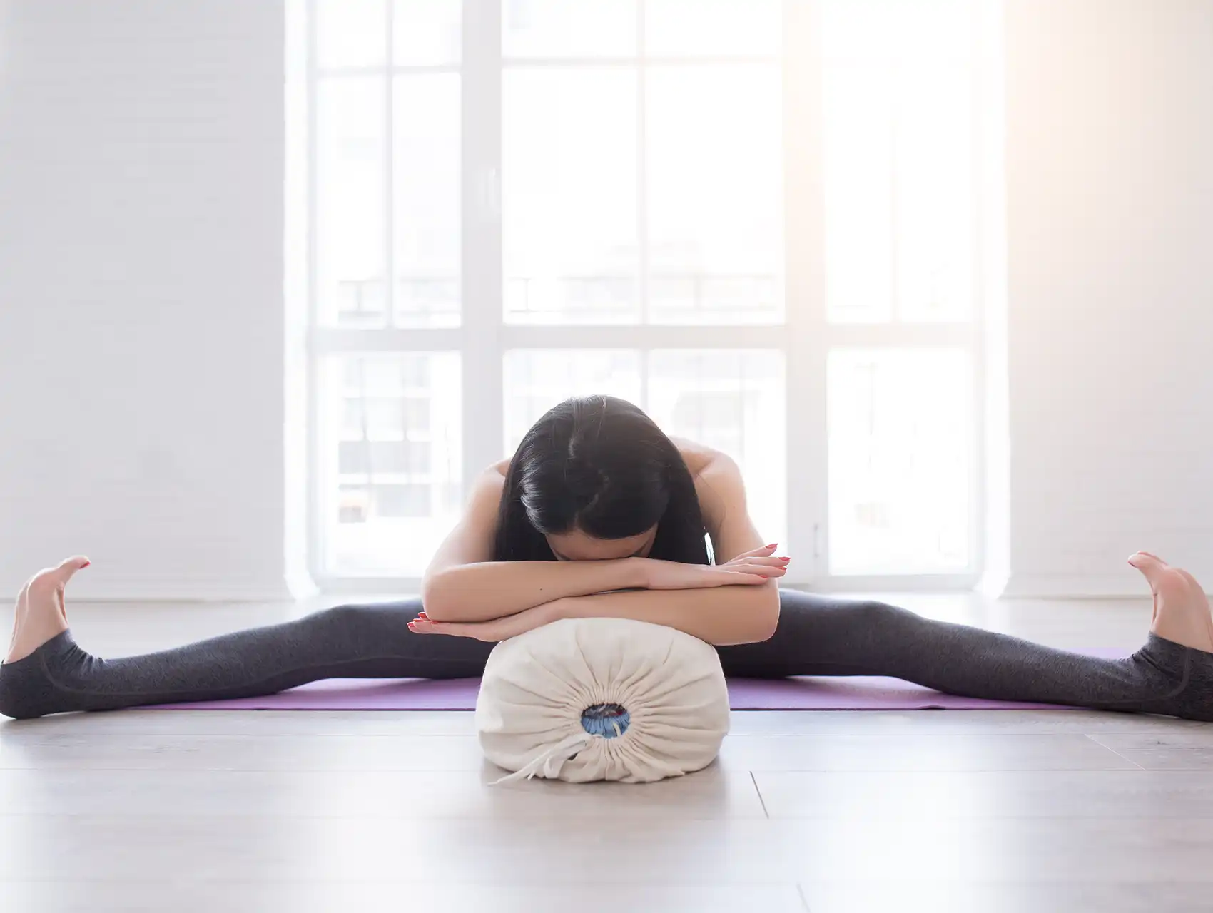 Woman in wide-legged forward bend resting on white yoga bolster with bolsters