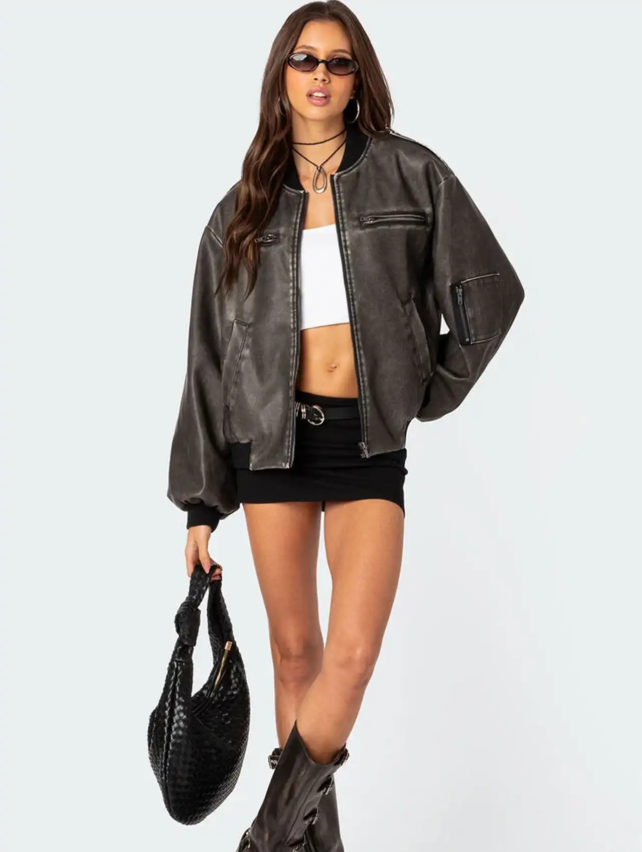 Woman wearing a brown bomber jacket with a cropped top and mini skirt, styled with boots and sunglasses.