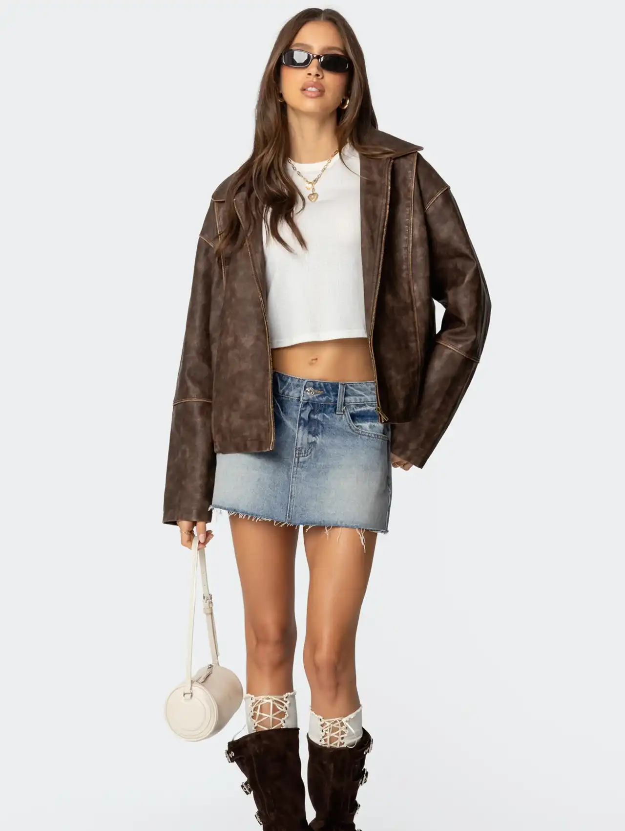 Woman wearing a brown bomber jacket with a denim skirt, white crop top, and accessories.