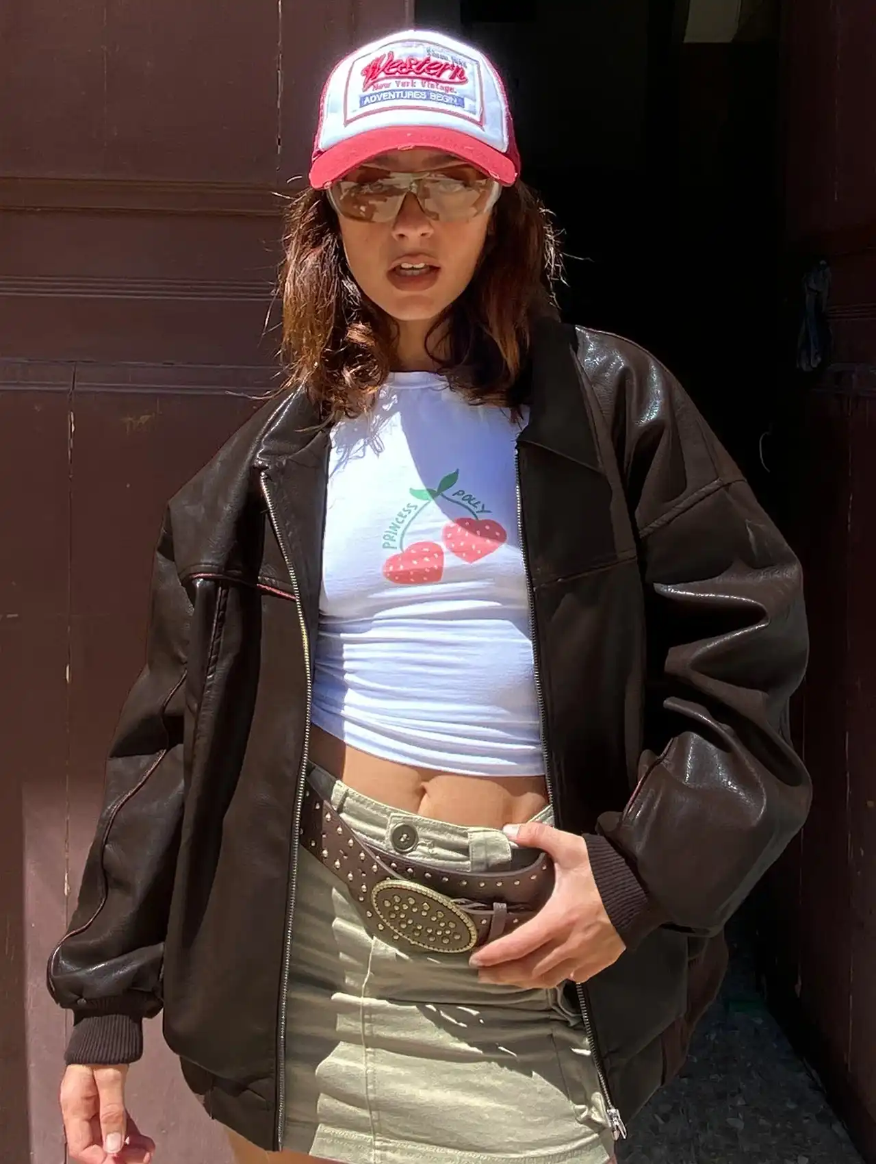 Model in a brown bomber jacket styled with a cap, mini skirt, and graphic tee for a casual street look.