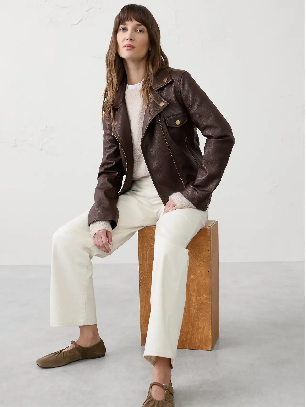 Woman seated in a brown bomber jacket styled with cream pants and a light sweater.