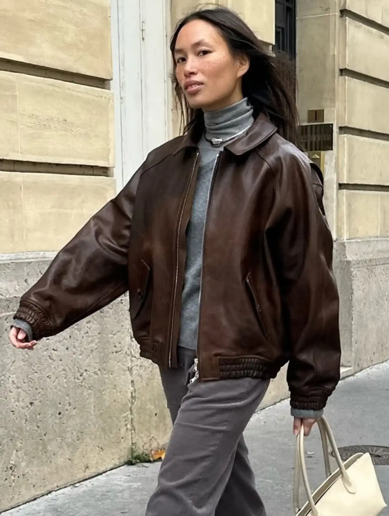 Woman walking in a brown bomber jacket layered over a gray turtleneck and trousers.