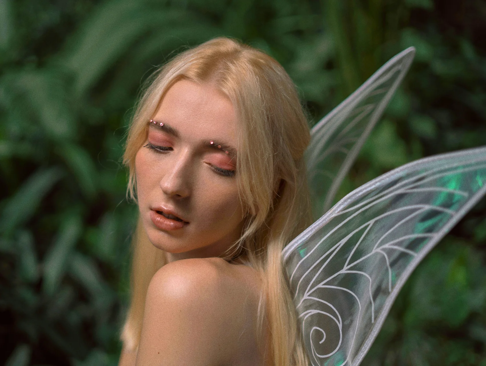 Fairy Halloween makeup look with rosy pink eyeshadow, crystals along the brows, glossy lips, and sheer iridescent wings