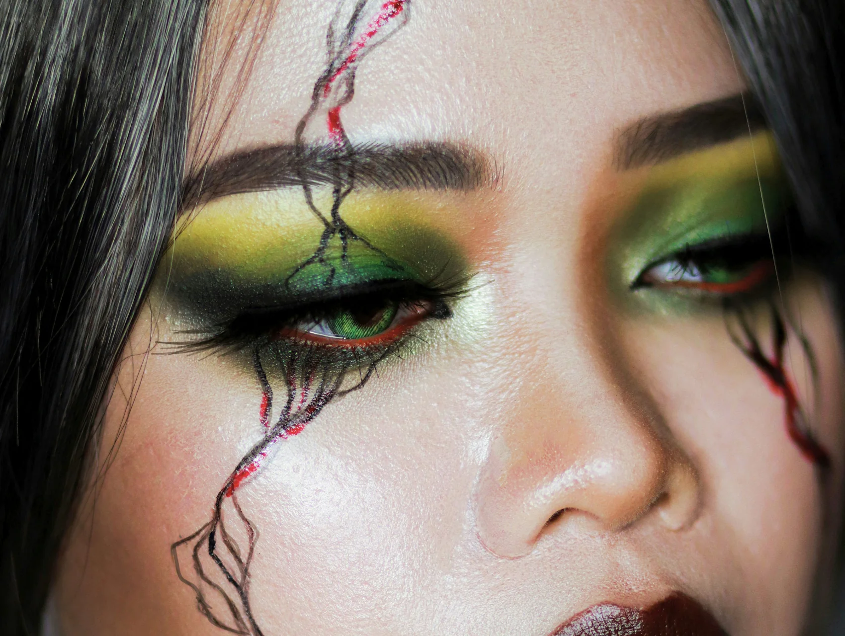 Zombie Halloween makeup look with green and yellow eyeshadow, red waterline, and black and red veins running down the cheek