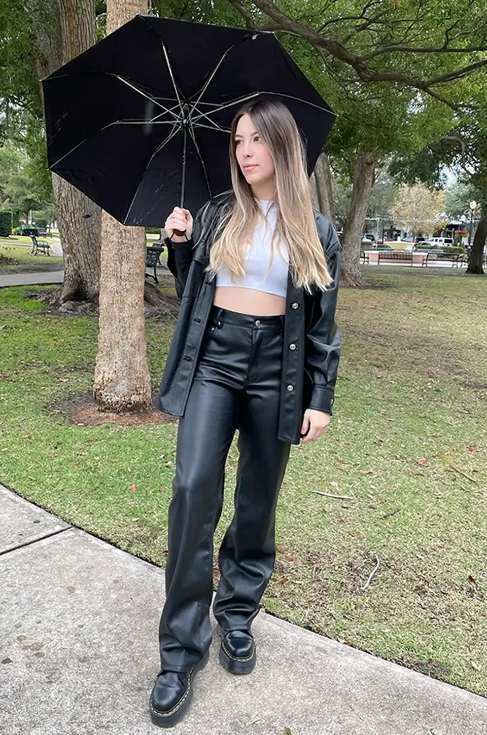 Woman wearing black faux leather pants with a cropped top and leather jacket, holding an umbrella in a city park.