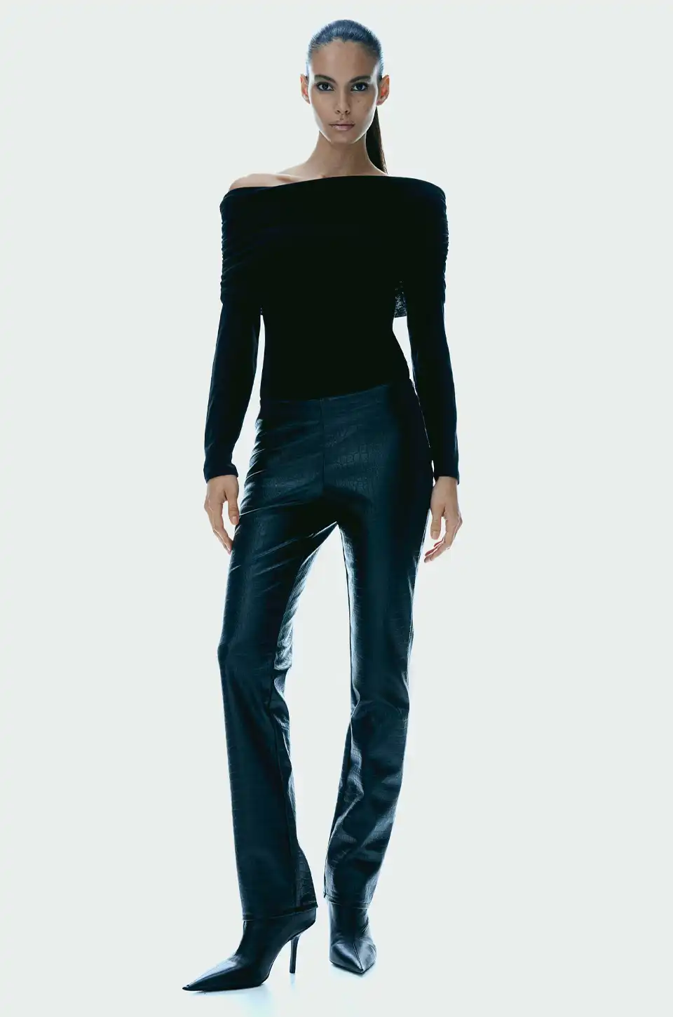 Model in slim black faux leather pants paired with an off-shoulder black top and stiletto boots.