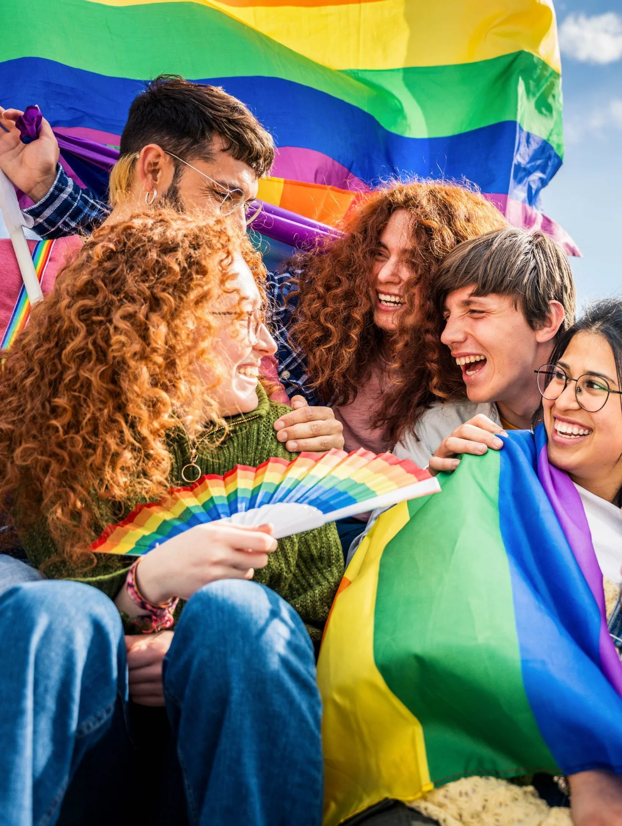 Group of LGBTQ+ friends laughing together with rainbow pride flags and fans during Pride Month celebrations