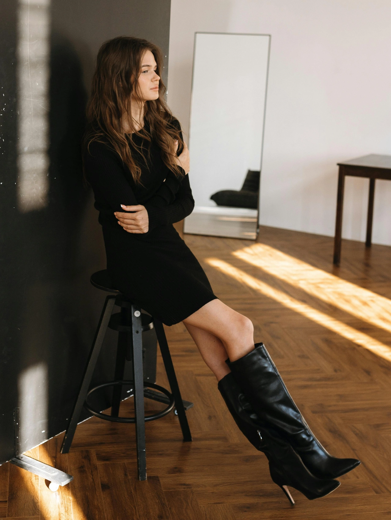 Woman sitting on a stool wearing a chic black outfit with stylish knee-high black boots – black boots fall outfit ideas.
