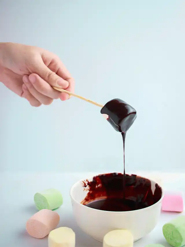 a hand holding a stick with chocolate on a stick over a bowl of liquid