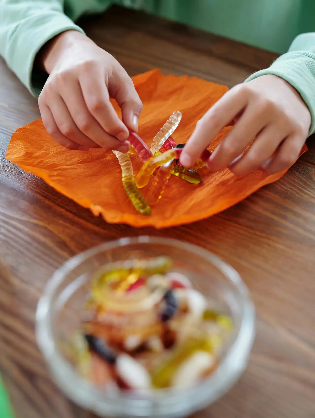 Child holding gummy worms on orange paper, vegan halloween candy option.