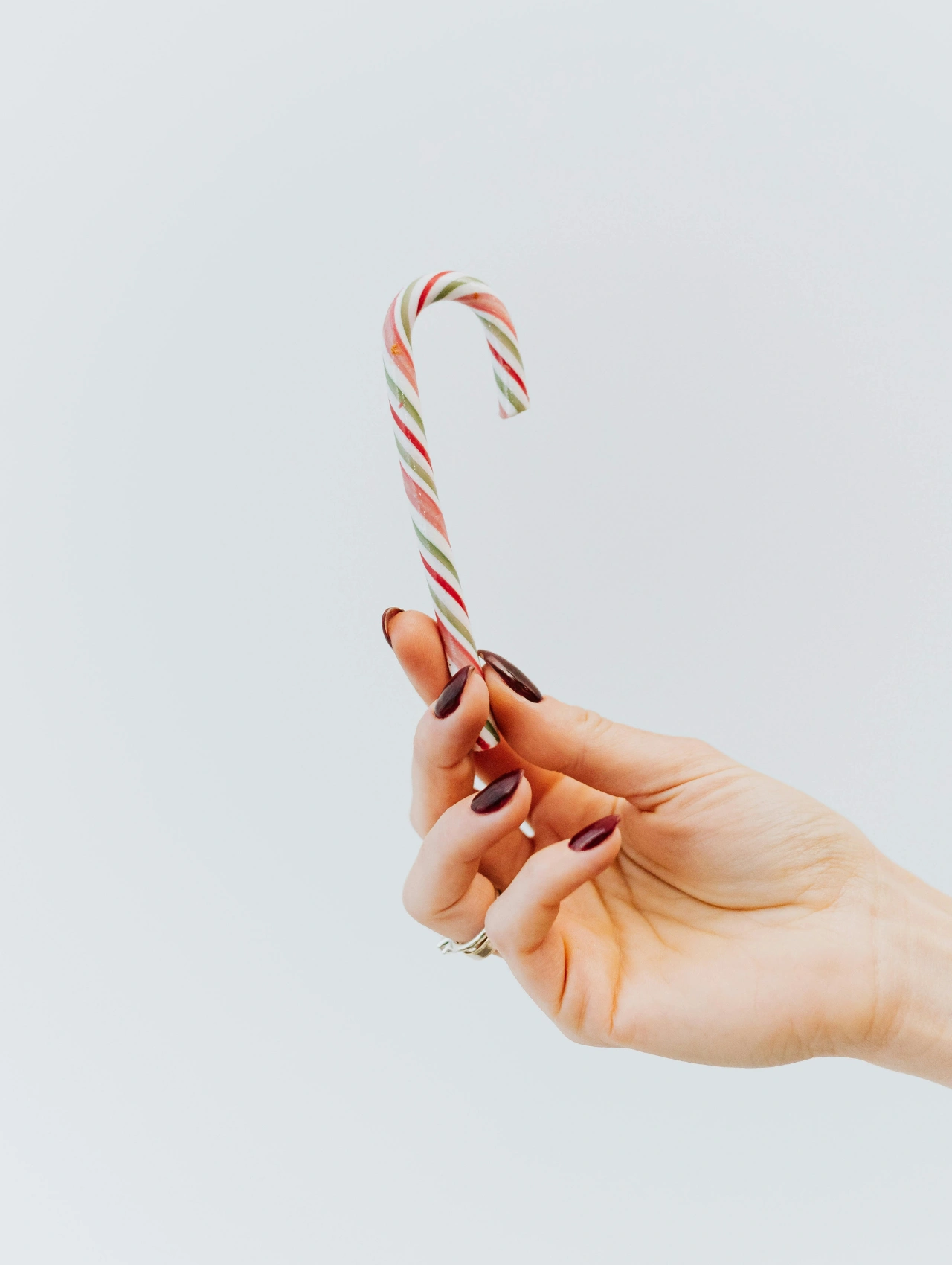 Hand holding a candy cane while showing deep burgundy christmas gel nails with a clean, minimal background.