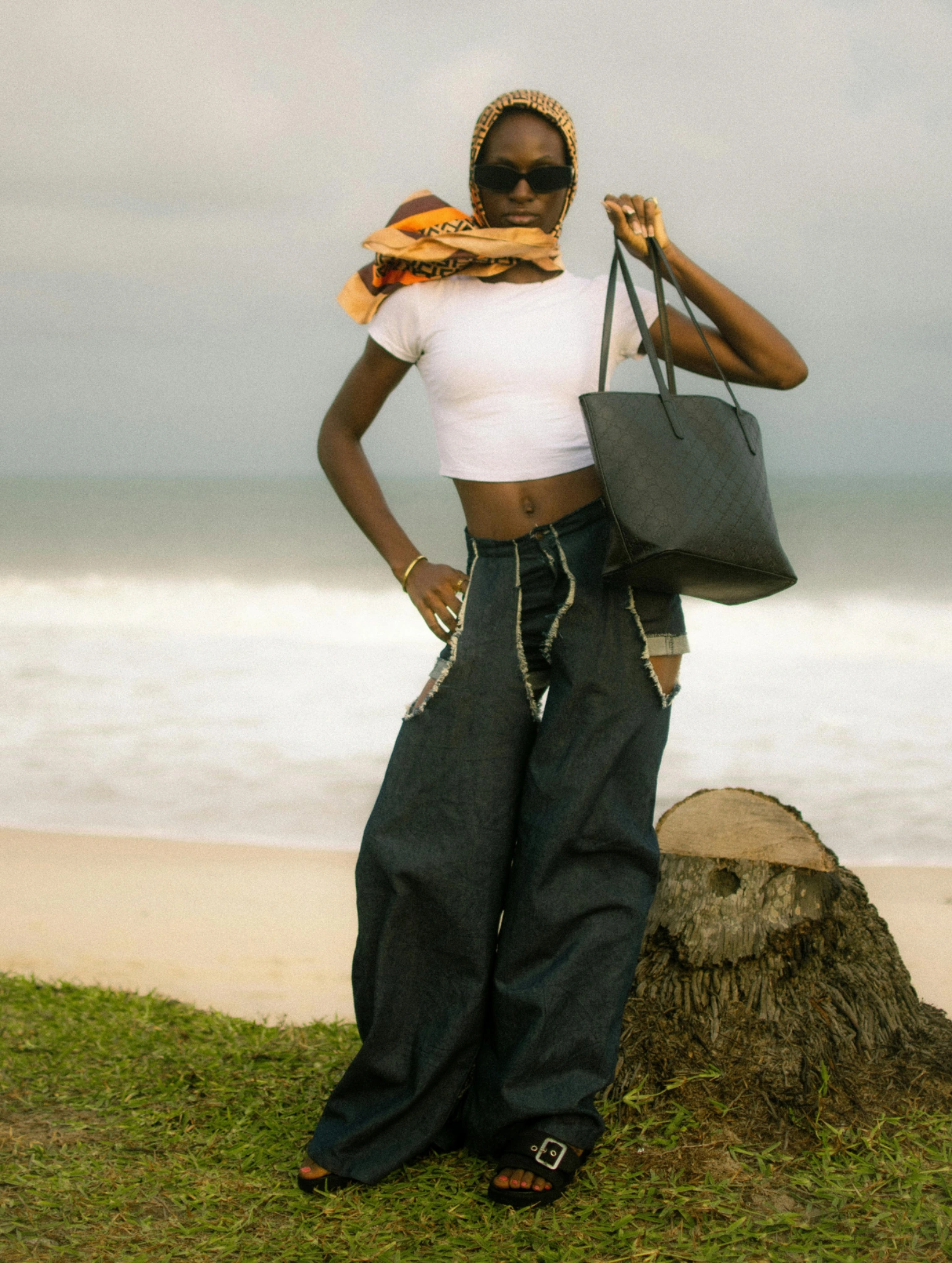 Quirky Spring Fashion Trends, stylish woman posing by the beach in oversized dark denim pants, white crop top, headscarf, sunglasses, and black tote bag, showcasing bold spring fashion trends.
