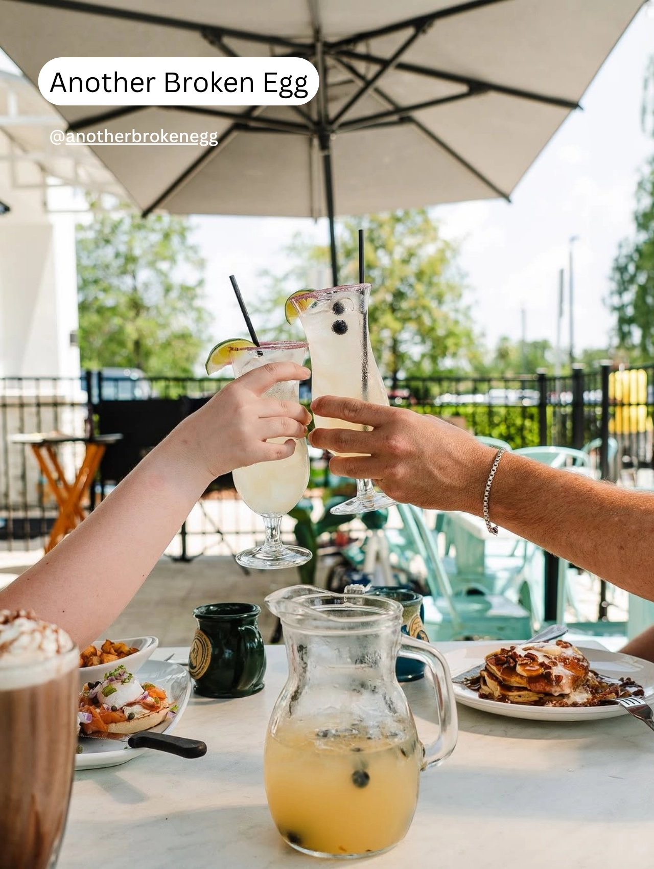 Two people clinking cocktails at an outdoor brunch table at Another Broken Egg, a sunny pet friendly restaurant in Orlando.