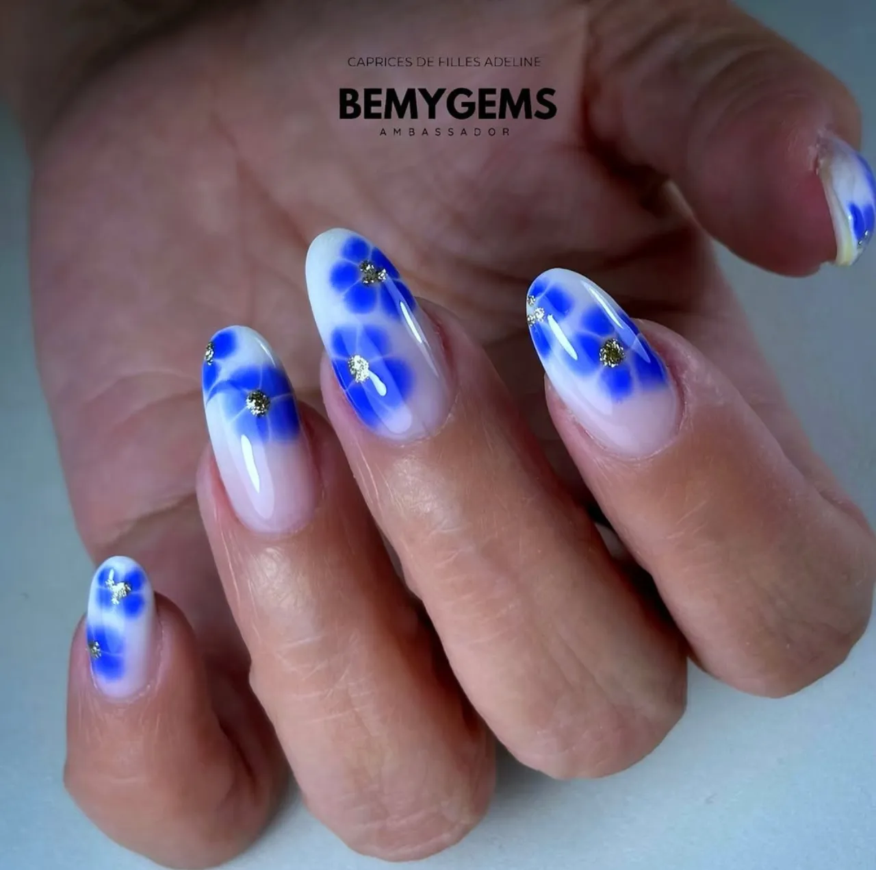 Diseños de uñas acrílicas con flores azules difuminadas y centros brillantes de glitter dorado.