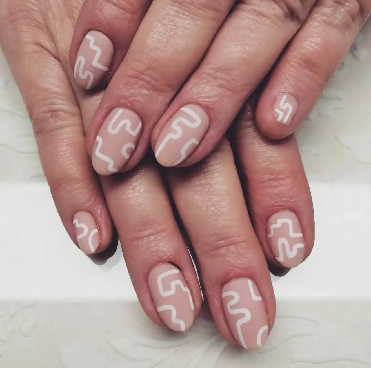 Diseños de uñas acrílicas cortas con líneas abstractas blancas sobre base nude.
