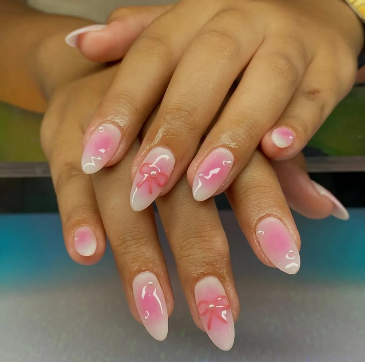 Efecto aura en uñas coquette con lazo rosa, detalles brillantes y acabado romántico.