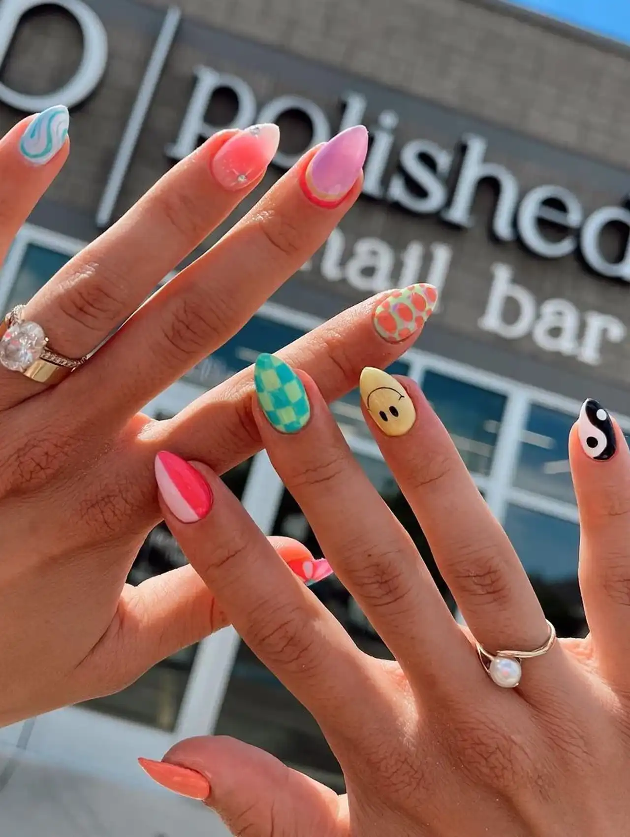 Two hands with colorful and funky summer nail designs, including smiley face, yin-yang, checkerboard, and swirls