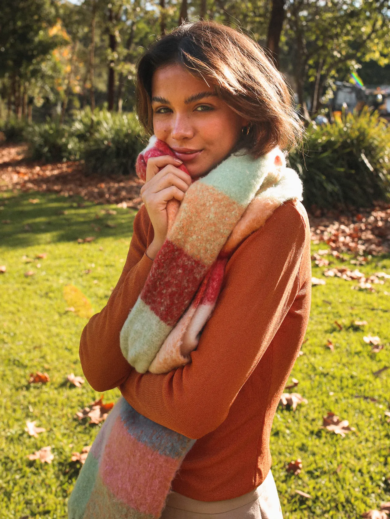 Smiling woman in a rust knit top and chunky plaid scarf on an autumn lawn, styling warm sweaters for women