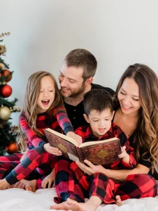 Familia leyendo juntos en pijamas de Navidad rojas y negras junto al árbol decorado, reflejando la unión y alegría de las fiestas.
