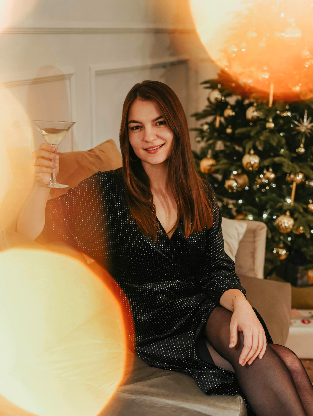 Outfit para año nuevo con vestido negro elegante y copa en mano frente a un árbol navideño decorado.
