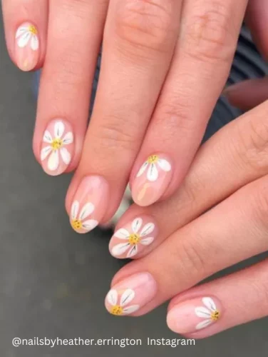 Close-up of a hand displaying nails painted with a soft pink base and adorned with intricate white daisy patterns, each with a yellow center.