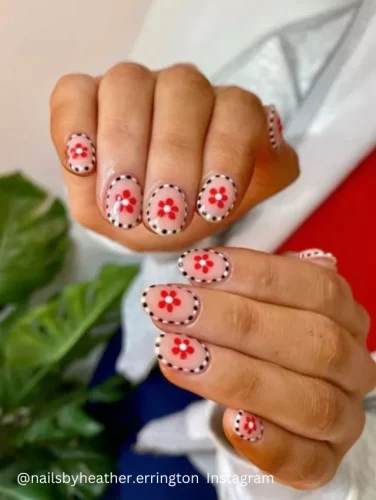 Close-up of hands displaying nails with a unique design featuring red floral patterns and black polka dots over a transparent base.