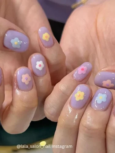 Close-up of hands displaying nails painted in a soothing lavender shade, each adorned with small, simple flower accents in varied pastel colors.