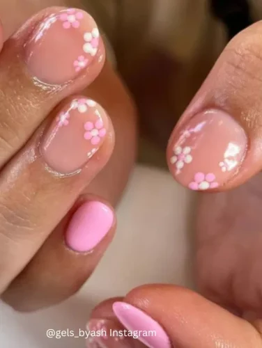 Close-up of hands displaying nails painted in a soft pink hue, accented with delicate white floral patterns near the cuticles.