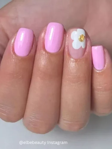 Close-up of hands displaying nails painted in a vibrant pink shade, featuring one accent nail with a white floral design and a touch of gold leaf.