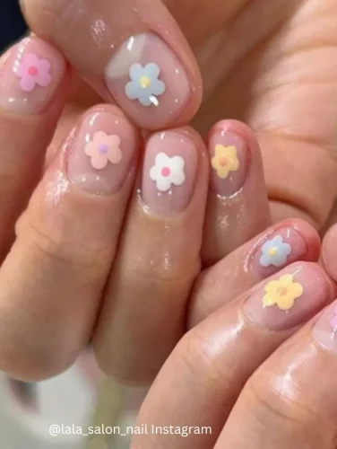 Close-up of hands displaying nails painted with a clear glossy base, each adorned with small pastel-colored flower decals.