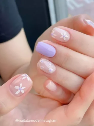 Close-up of hands displaying nails painted with alternating clear and soft lilac backgrounds, each adorned with simple white floral patterns.