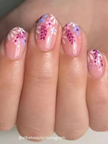 Close-up of hands displaying nails painted with a clear pink base, each detailed with intricate lilac and white floral patterns.