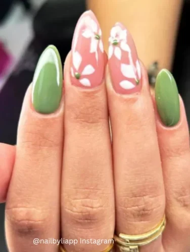 Close-up of hands displaying nails painted in alternating shades of soft pink and rich matcha green, with intricate white floral patterns on the pink nails.