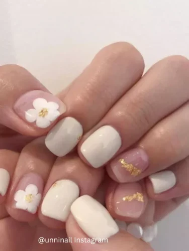 Close-up of hands displaying nails painted in a creamy white hue, some adorned with subtle floral designs and small gold leaf accents.