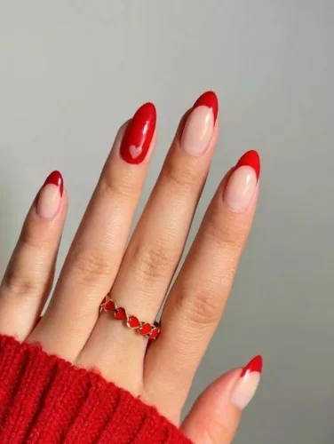 A timeless Valentine's nail design featuring a soft nude base with bold red French tips. A subtle heart accent on the ring finger adds a romantic touch, making this style perfect for those who love classic yet playful designs. The almond shape enhances elegance, while the deep red shade complements the warm and cozy vibes of the season.