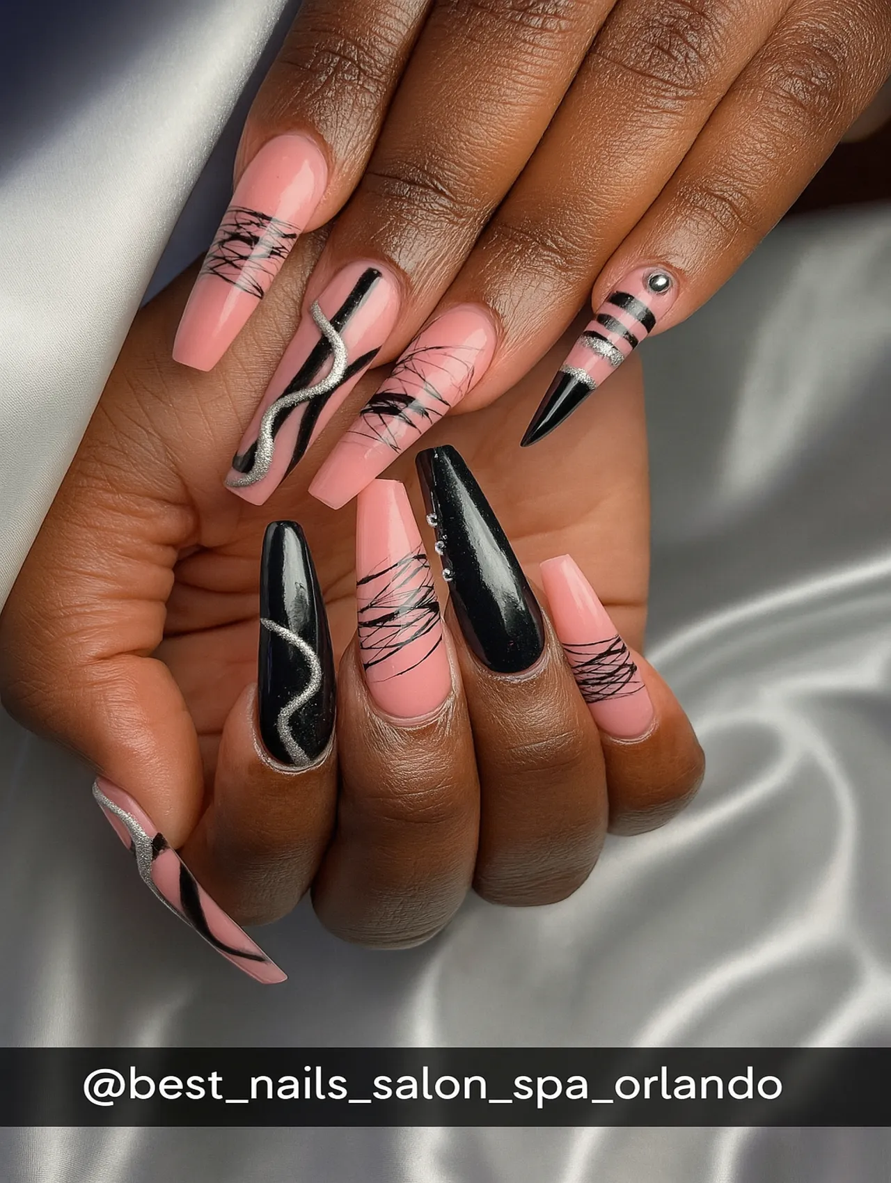 Close-up of coffin-shaped nails featuring pink base polish with black geometric patterns, silver glitter swirls, and studded accents.