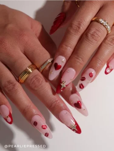 Close-up of a hand showcasing Valentine’s Day-themed nail art, featuring long, stiletto nails with designs of hearts, lip prints, and golden stars against a sheer pink background.