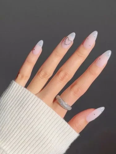 Este elegante y moderno diseño de uñas presenta un degradado de rosa suave a gris, culminando en una punta chic y afilada. Cada uña está sutilmente adornada con pequeños y brillantes diamantes de imitación cerca de la cutícula, añadiendo justo la cantidad adecuada de brillo. Una sola uña presume de un delicado patrón de corazón formado por pequeños puntos, realzando su encanto minimalista. Este diseño es perfecto para aquellos que aprecian la elegancia discreta con un giro contemporáneo, convirtiéndolo en una elección elegante para el Día de San Valentín.