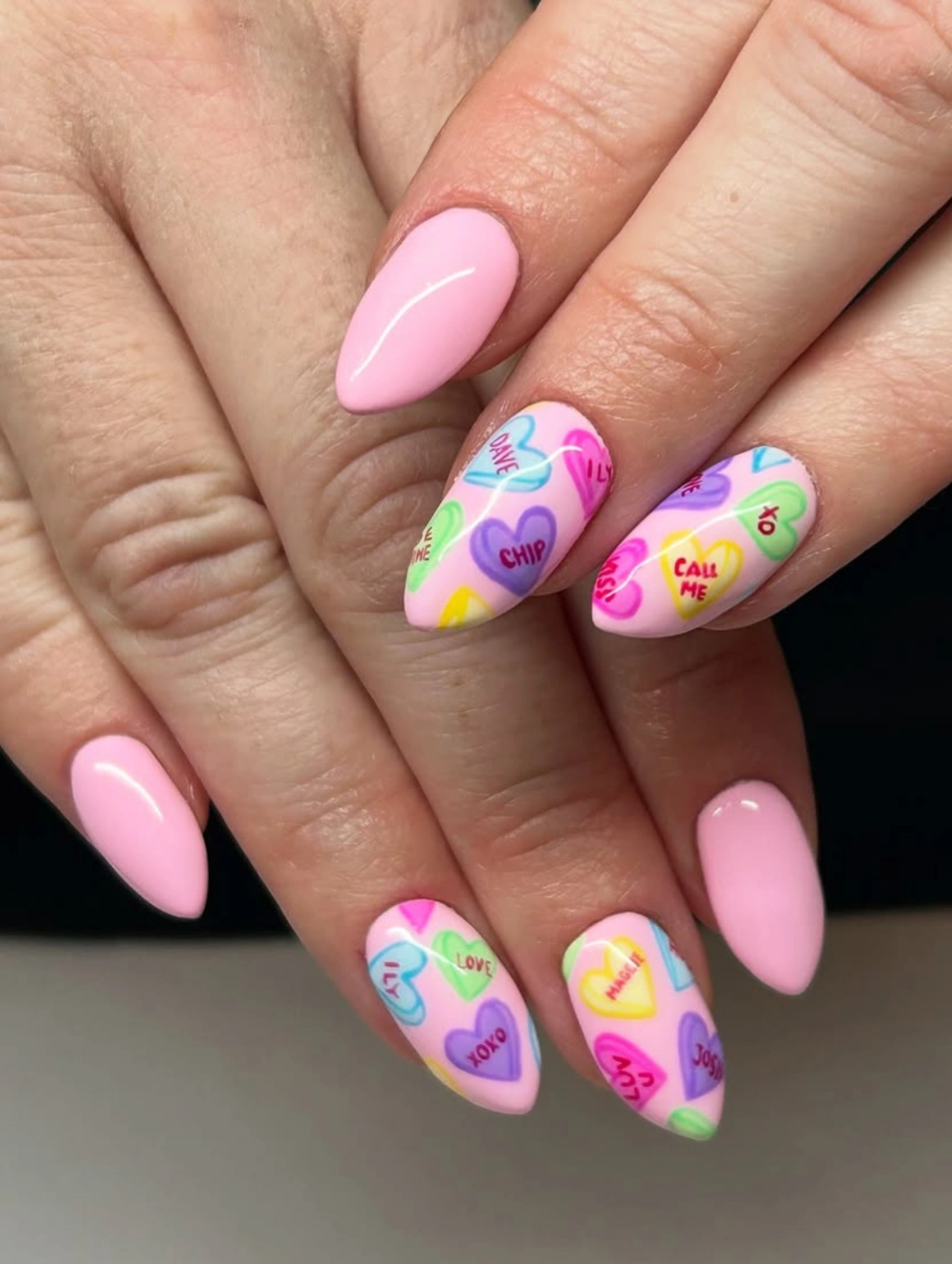 Candy Heart Conversation Valentineโs Nails Almond with almond-shaped pink nails featuring colorful heart messages.