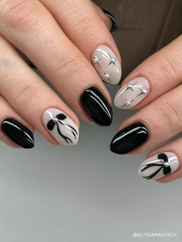 A sophisticated black and nude nail design featuring glossy black polish, delicate bow accents, and silver embellishments.