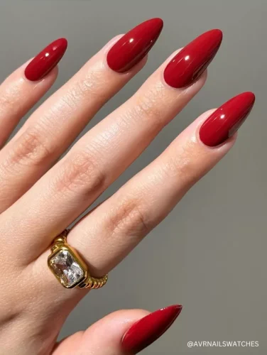 A hand with long, almond-shaped nails painted in a glossy, deep red polish, complemented by a gold ring with a large gemstone.