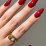 A hand with long, almond-shaped nails painted in a glossy, deep red polish, complemented by a gold ring with a large gemstone.