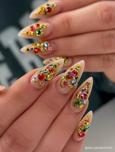 A set of long stiletto nails with a nude base adorned with multicolored rhinestones and intricate gold embellishments.