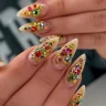 A set of long stiletto nails with a nude base adorned with multicolored rhinestones and intricate gold embellishments.