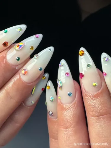 Elegant stiletto nails with a sheer milky base adorned with scattered multicolored rhinestones for a delicate yet glamorous look.